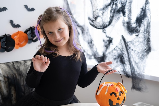 little girl in costume of witch holding pumpkin jack with candies, celebrating Halloween at home