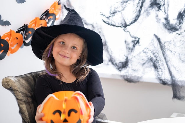 little girl in costume of witch holding pumpkin jack with candies, celebrating Halloween at home