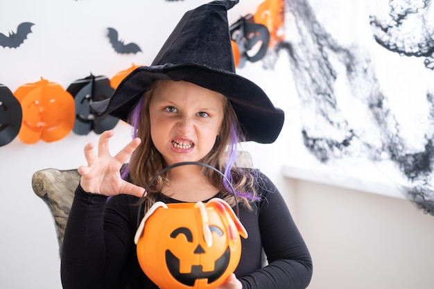 little girl in costume of witch holding pumpkin jack with candies, celebrating Halloween at home