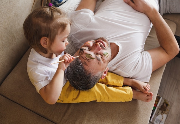 A little girl on the couch carefully paints her father's face during family games together