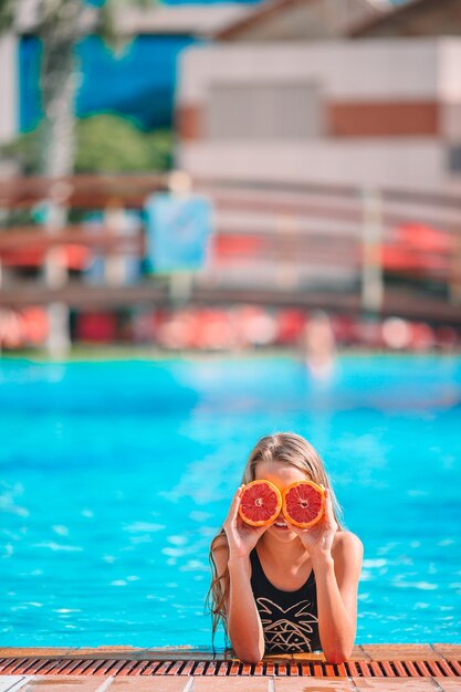 Little girl covering eyes with orange halves near eyes on background swimming pool