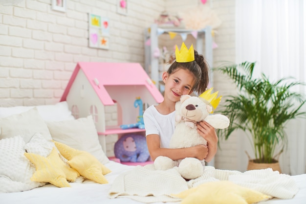 Bambina in corona, seduto sul letto nella stanza dei bambini e abbracciando orsacchiotto