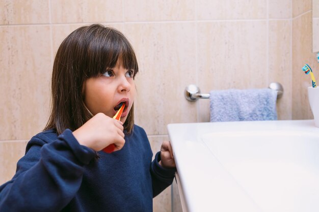 Little girl in dark blue sweatshirt, brushing her teeth while looking in the mirror, in the bathroom. Tooth brushing, hygiene and childhood concept.