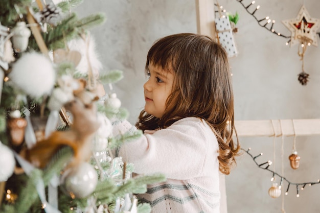 Little girl decorates the Christmas tree with toys. A cute baby is preparing at home for the celebration of Christmas.