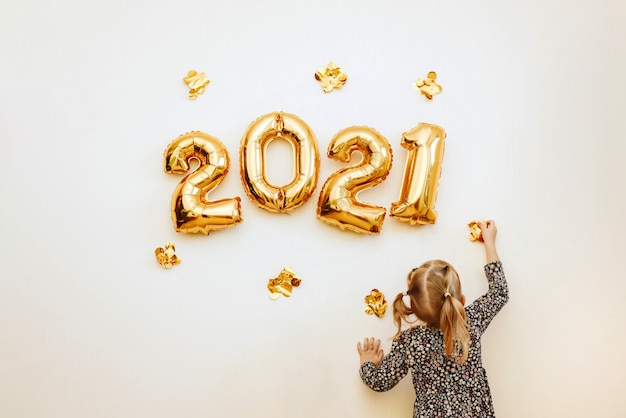 Little girl decorates the wall of the house with golden numbers 2021. Preparing the house for Christmas