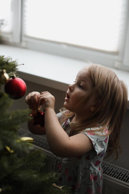 Little girl decorating christmas tree with toys and baubles Cute kid preparing home for xmas celebration