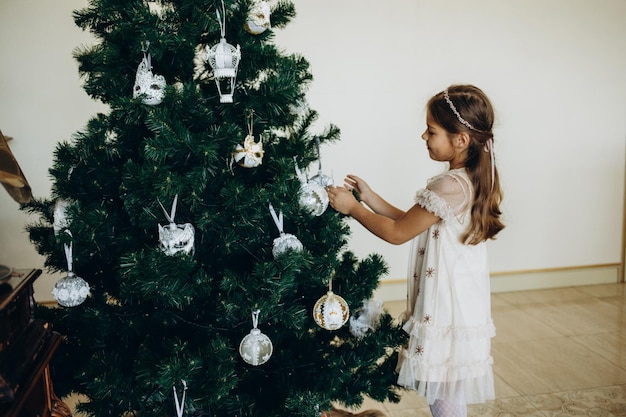 Little girl decorating christmas tree with toys and baubles Cute kid preparing home for xmas celebration