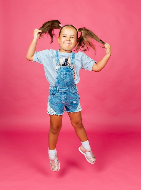 Bambina in salopette di jeans che salta su uno sfondo rosa. bambino allegro. figli.