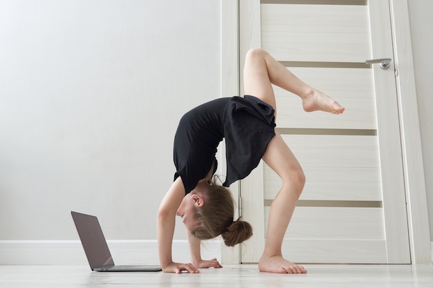 Little girl doing gymnastics exercises at home using online learning with laptop computer