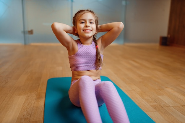 Маленькая девочка делает упражнения пресса на циновке в тренажерном зале, фитнес-тренировки. Девушка в спортивной одежде, ребенок на тренировке в спортивном клубе