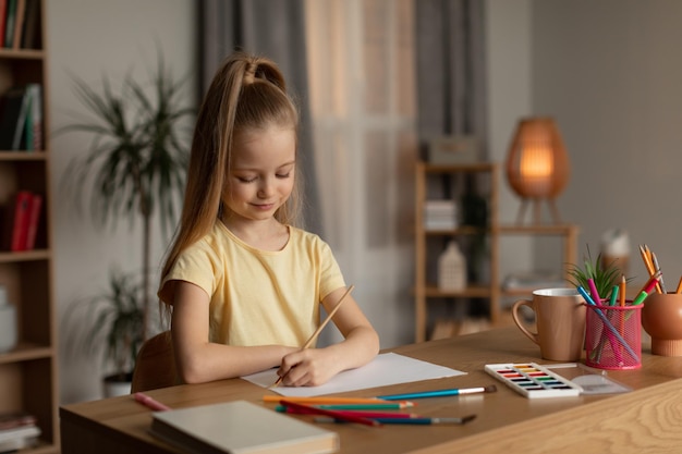 Little Girl Drawing With Paintbrush Sitting At Desk At Home