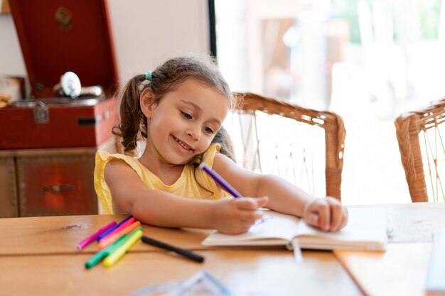 A little girl draws in a notebook with colored markers, sitting at a wooden table enjoys her creative process.