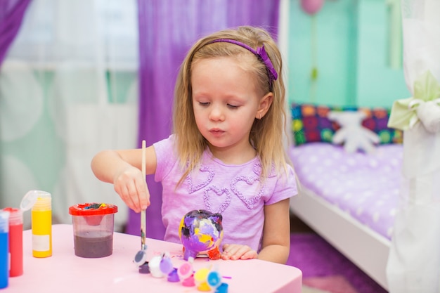 Маленькая девочка рисует красками на своем столе в комнате