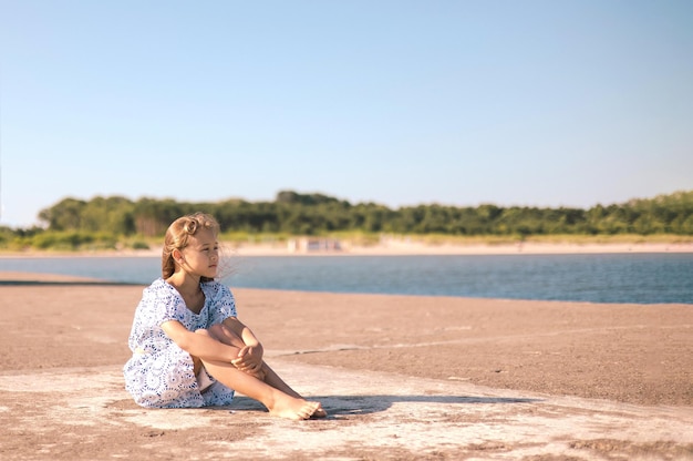 little girl in a dress on the beach with developing long blond hair in the wind and looking on sea