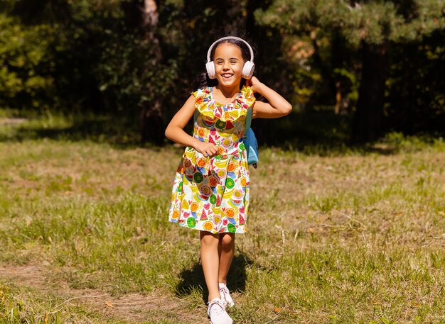 Little girl in a dress and headphones is having fun and jumping in the park on the grass.