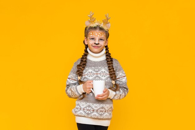 A little girl dressed as a christmas reindeer holds a cup of hot cocoa in her hands a child in a sweater is enjoying the holiday yellow isolated background