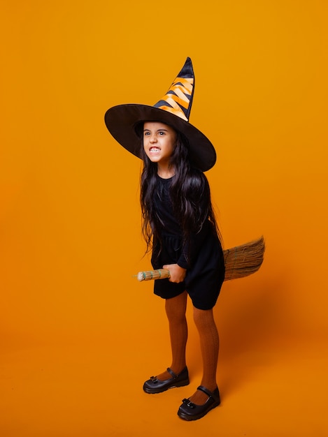 Little girl dressed as a Halloween witch in a black dress and hat flies on a broomstick on a yellow background.