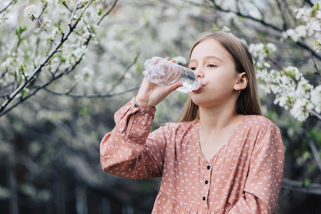 Una bambina beve acqua dolce pulita da una bottiglia di plastica vicino a un albero in fiore nel giardino.