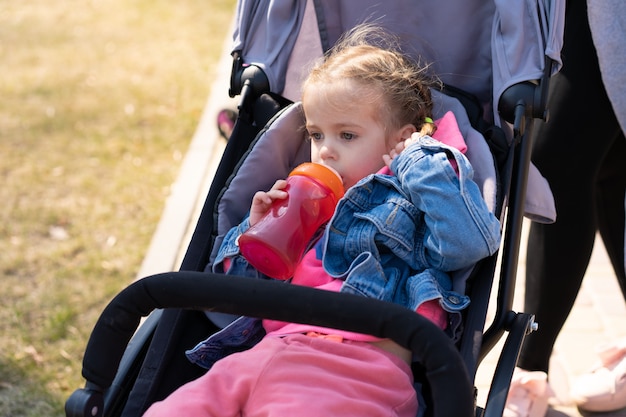 Little girl drinks juice from a bottle while sitting in a baby carriage on a walking