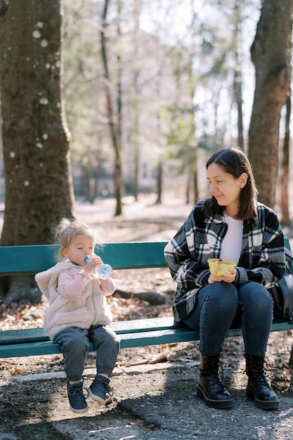 Little girl drinks water from a bottle while sitting next to her mother on a bench in a sunny park