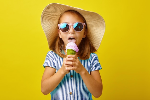 Фото Маленькая девочка ест мороженое на изолированном желтом фоне, одетая в летнюю одежду и солнцезащитные очки