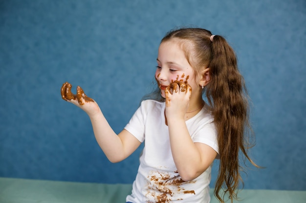 어린 소녀는 초콜릿을 먹고 흰색 티셔츠, 얼굴, 손을 문지릅니다.