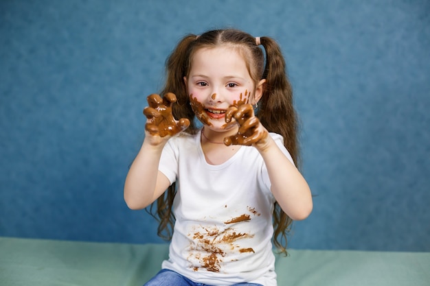 小さな女の子はチョコレートを食べ、彼女の白いTシャツを汚し、顔をして彼に手渡します