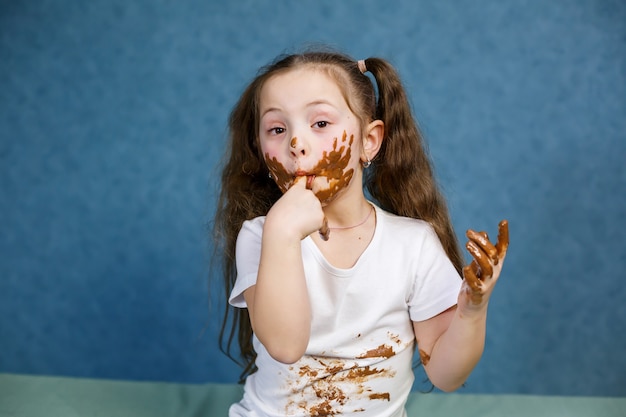 小さな女の子はチョコレートを食べ、彼女の白いTシャツを汚し、顔をして彼に手渡します