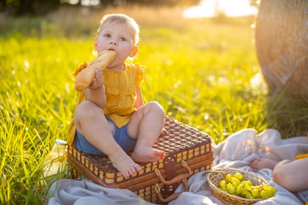 Little girl eats fresh baguette and fruit on a picnic at sunset lights in nature