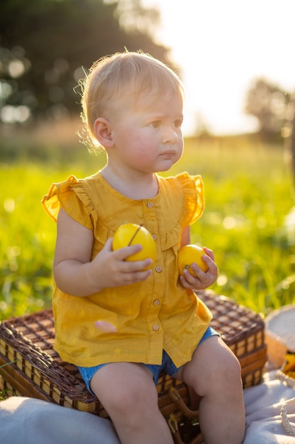 Little girl eats fresh fruits on a picnic at sunset lights in nature