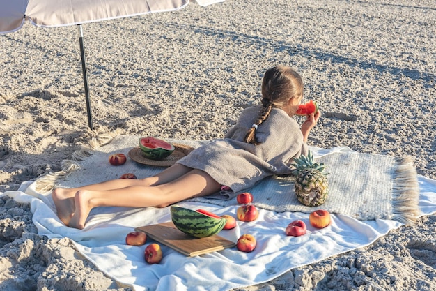 ビーチの毛布の上に横たわっている小さな女の子が果物を食べている