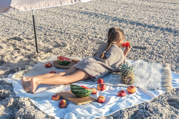 少女はビーチで毛布の上に横たわって果物を食べる