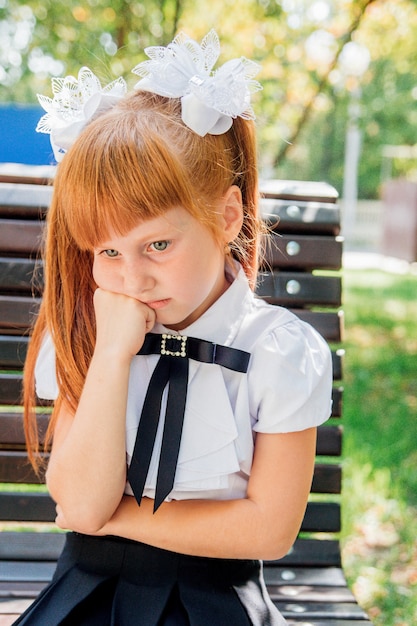 A little girl, an elementary school student, is sitting on a bench on the street, a schoolgirl has an offended and sad expression on her face.