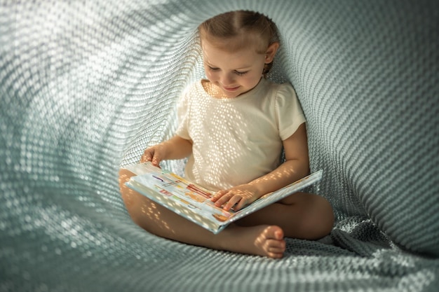 Little girl enjoying of review a book under blue knitted plaid in sunny morning