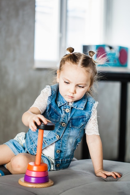 Foto bambina di aspetto europeo seduto sul divano e giocando con una piramide, giocattolo di legno per lo sviluppo del bambino