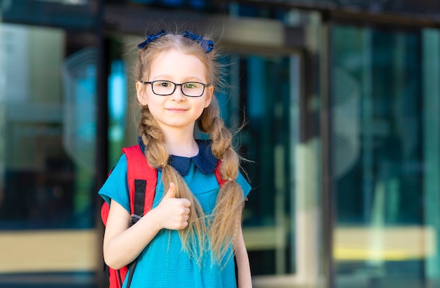 小さな女の子の眼鏡一年生。幸せな子供が学校に戻って、親指を立てるジェスチャー。ランドセルを持つ子供