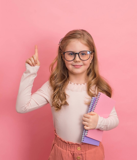 Photo little girl in eyeglasses with copybooks on pink background education concept