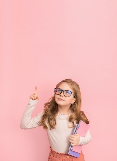 Photo little girl in eyeglasses with notebooks or copybooks on pink background pointing to copyspace