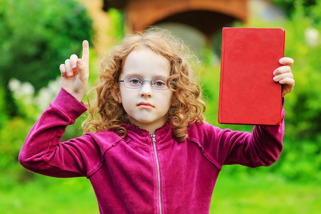 Little girl in eyeglasses with red book and finger up.