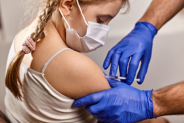 의사의 사무실에서 얼굴 마스크에 어린 소녀가 예방 접종을받습니다. COVID-19 코로나 바이러스, 독감에 대한 백신 주사기. 약.