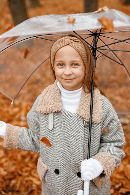 Little girl in fashion clothes standing in autumn forest girl\
posing for a photo and holding a transparent umbrella girl wearing\
grey coat and a headband