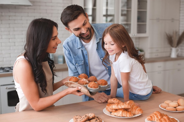 Little girl and father sitting on the kitchen table and looking at mather holding freshly baked muffins