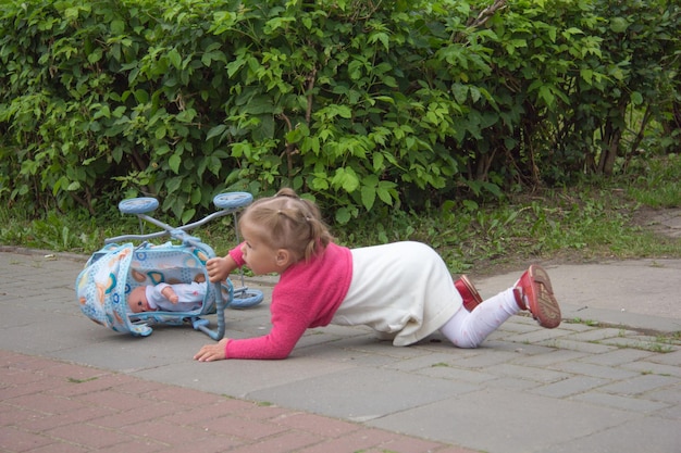 小さな女の子が夏の公園でおもちゃのベビーカーで倒れた