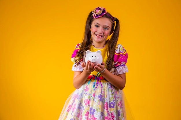 Little girl in festa junina clothes holding a piggy bank on yellow background Finance and festa junina concept