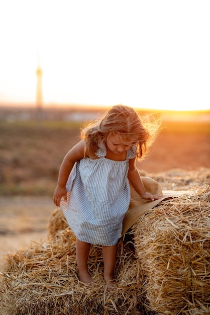 Little girl in a field with hay rolls at sunset Landscape of a field with haystacks of straw at sunset A little girl in a blue dress stands in the middle of a meadow Harmony and love of nature