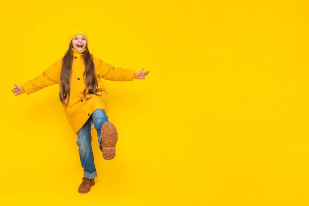 A little girl in full height in an autumn coat down jackets and hats rejoices in autumn the child joyfully raises his leg with his arms outstretched to the sides yellow isolated background