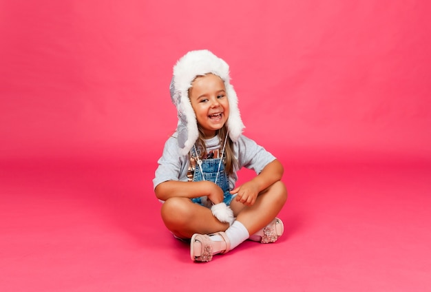 Little girl in a fur winter hat sits on a pink background. Winter fashion warm clothes concept. Children's warm woolen hat