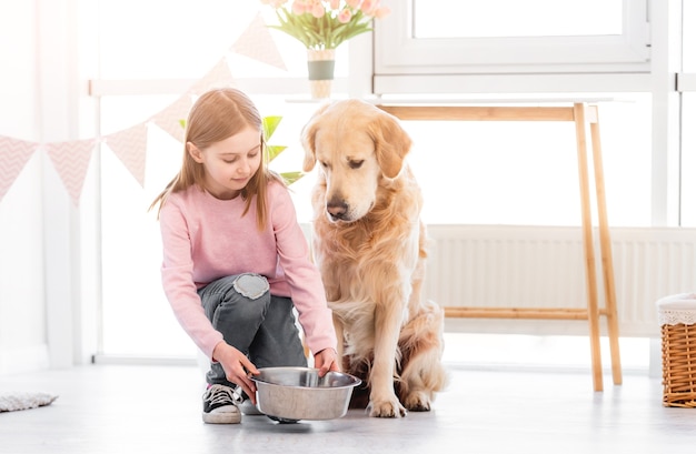 Маленькая девочка дает милой собаке золотистого ретривера металлическую миску с кормом в солнечной комнате дома