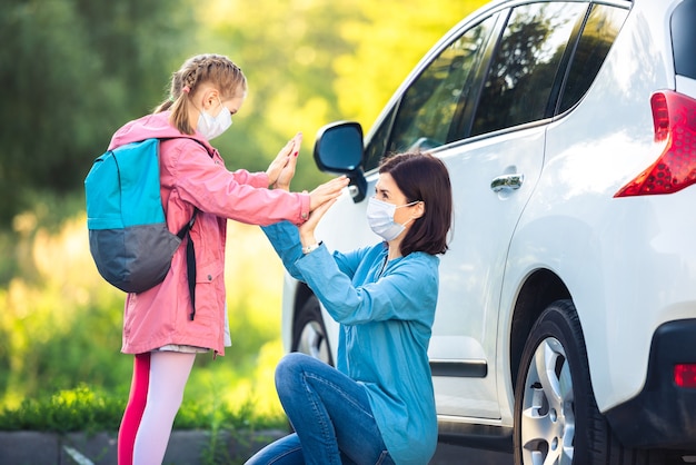 Маленькая девочка дает матери пять после школы возле машины в медицинских масках