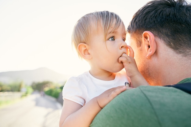 Little girl gnaws fingers while sitting in the hands of dad